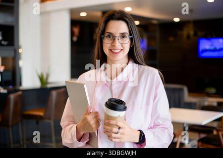Donna sorridente con occhiali che reggono un laptop e una tazza di caffè in un ambiente interno moderno, ritratto informale di uno studente o di un freelance in un ambiente luminoso Foto Stock