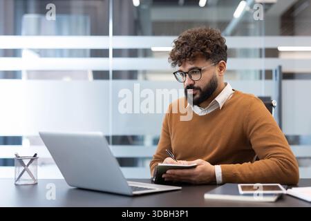 Giovane uomo con capelli ricci e barba che indossa occhiali, concentrato sull'apprendimento online dal suo portatile, prendendo appunti in un notebook presso una scrivania in un moderno spazio di lavoro aziendale Foto Stock