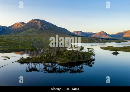Una tranquilla scena lacustre caratterizzata da una piccola isola piena di alberi, acque calme e montagne lontane immerse nella calda luce dorata. Galway, Irlanda Foto Stock