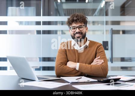 Uomo d'affari sicuro che indossa occhiali da vista e un maglione marrone seduto a una scrivania in un ufficio moderno, sorride e guarda la fotocamera, proiettando professionalità e successo Foto Stock