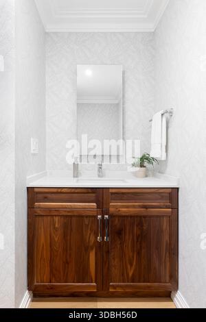 Un elegante lavandino per toilette con un armadietto in legno scuro con ripiano bianco, rubinetto moderno e carta da parati bianca e grigia. Foto Stock
