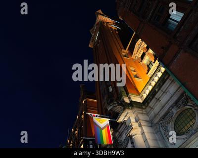 Manchester, Regno Unito - 21 novembre 2025: Un'alta torre di mattoni a Manchester si illumina di notte, con una bandiera arcobaleno che pende dalla facciata. Foto Stock