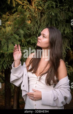 La giovane donna si trova vicino ai rami sempreverdi, toccando delicatamente gli aghi di pino, luce del giorno soffusa, toni verdi profondi, umore contemplativo Foto Stock