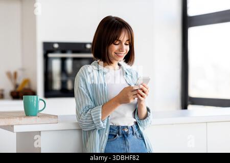 Una giovane donna ama controllare il suo telefono mentre si trova in una cucina moderna con una tazza di caffè Foto Stock