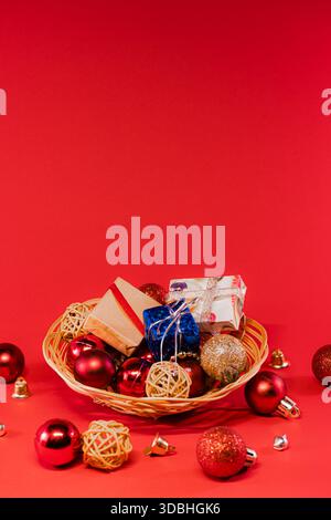Una disposizione festosa di ornamenti natalizi in tessuto rosso, oro e naturale in un cestino di vimini, su uno sfondo rosso brillante. Lucente e scintillante Foto Stock