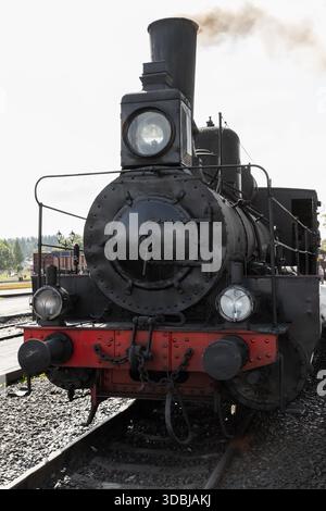 Una storica locomotiva a vapore si trova di fronte alla telecamera su un binario ferroviario, la sua grande caldaia rivettata e il fronte rosso brillante contrastano con il fumo e il cielo limpido Foto Stock