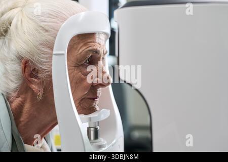Senior donna caucasica sottoposta a esame oculare utilizzando oftalmologia macchina diagnostica in clinica medica, profilo laterale che mostra paziente che si concentra sulla procedura di test di visione Foto Stock