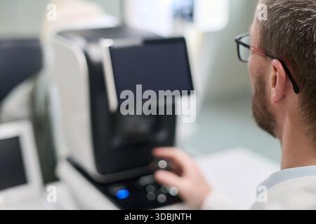 Uomo di mezza età caucasico che utilizza il dispositivo diagnostico oftalmico in ambiente clinico, utilizzando la manopola di controllo mentre è seduto di fronte all'apparecchiatura medica durante la procedura di esame oculare Foto Stock