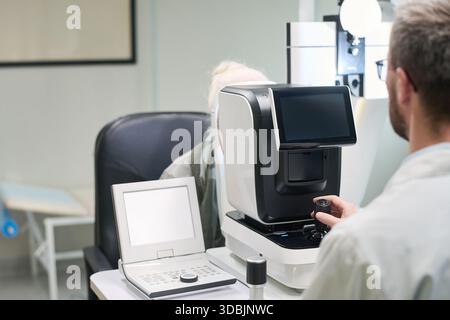 Donna anziana caucasica sottoposta a esame oculistico con autorefractor operata da uomo in studio medico, entrambi i soggetti si sono concentrati sulla procedura diagnostica, moderna apparecchiatura oftalmologica visibile Foto Stock