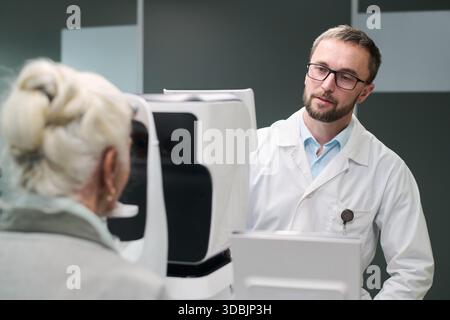 Donna anziana caucasica sottoposta a esame oculistico con dispositivo oftalmico mentre optometrista maschio adulto caucasico conduce test di visione in clinica medica moderna Foto Stock