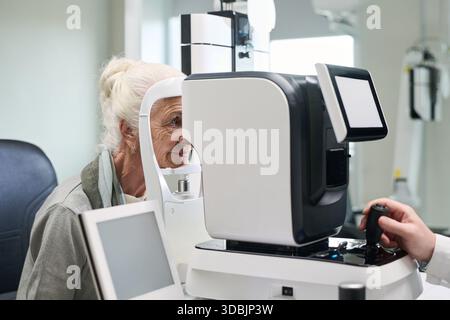 Donna caucasica anziana sottoposta a esame oculistico utilizzando apparecchiature oftalmoloiche avanzate, tecnico che opera macchina diagnostica in ambiente clinico, concentrandosi sulla salute della vista Foto Stock