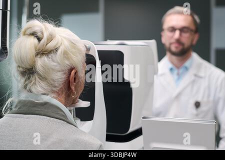 Donna caucasica anziana sottoposta a esame oculare utilizzando dispositivo oftalmico avanzato mentre optometrista maschio caucasico di mezza età supervisionando la procedura in clinica medica Foto Stock