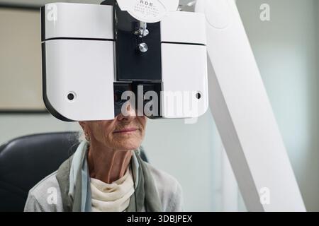 Donna caucasica anziana sottoposta a esame oculare utilizzando il phoropter presso l'ufficio optometrista, seduta su una sedia con il viso parzialmente visibile dietro il dispositivo diagnostico durante il test di visione Foto Stock