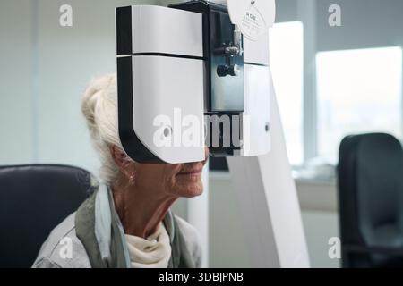 Donna caucasica anziana sottoposta a esame oculare utilizzando dispositivo oftalmologico in clinica medica, seduta con la testa posizionata contro la macchina durante il test di visione Foto Stock