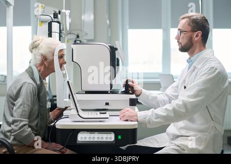 Donna caucasica anziana sottoposta a esame oculare con oftalmologo dispositivo medico operante in clinica, entrambi i soggetti si sono concentrati sulla procedura diagnostica, attrezzature moderne visibili Foto Stock