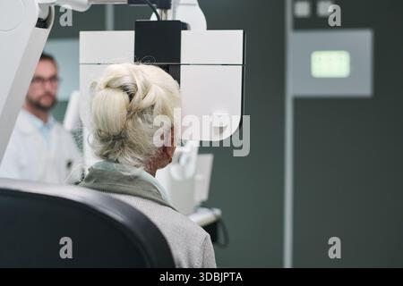 Donna caucasica anziana sottoposta a esame oculistico utilizzando la macchina diagnostica oftalmica in clinica medica, medico caucasico maschio osservando procedura in background, ambiente clinico visibile Foto Stock