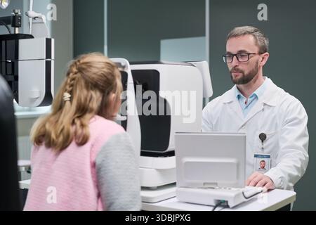 Uomo di mezza età caucasico optometrista che conduce l'esame oculare per la bambina caucasica utilizzando moderne attrezzature oftalmiche in clinica medica, entrambi concentrati sul processo diagnostico Foto Stock