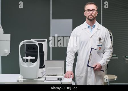 Ritratto di uomo caucasico di mezza età con barba che indossa un camice da laboratorio in piedi in una clinica di oftalmologia che tiene gli appunti accanto all'apparecchiatura diagnostica oftalmica, guardando la macchina fotografica Foto Stock