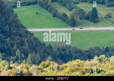 Vista aerea della strada di campagna che attraversa prati lussureggianti e colline boscose nella valle di montagna con due auto che guidano in direzioni opposte. Foto Stock