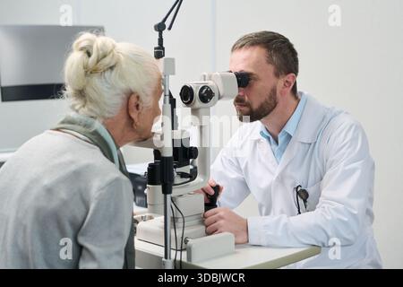 Donna anziana caucasica sottoposta a esame oculare da parte di optometrista maschio di mezza età caucasico utilizzando la lampada a fessura biomicroscopio in clinica medica, entrambi concentrati sulla procedura diagnostica Foto Stock