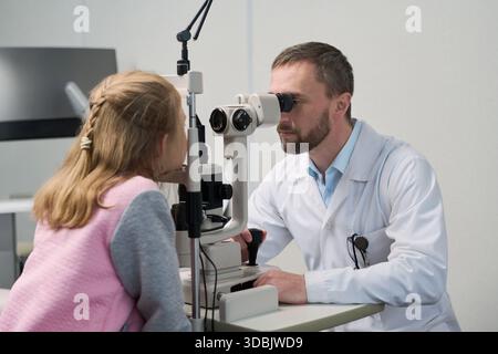 Uomo di mezza età caucasico che esegue l'esame oculare su bambino caucasico utilizzando apparecchiature oftalmologia in studio medico, entrambi concentrandosi sulla procedura diagnostica, ambiente clinico visibile Foto Stock