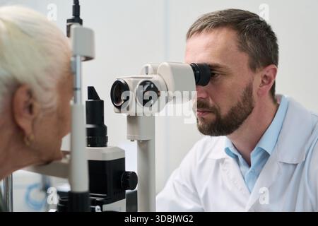 Uomo di mezza età caucasico che esegue l'esame oculistico su donna caucasica anziana utilizzando attrezzature oftalmologia in clinica medica, concentrandosi su pazienti valutazione della salute degli occhi Foto Stock