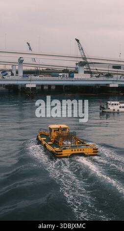 Imbarcazione industriale gialla che opera su un fiume urbano vicino a un ponte autostradale e all'infrastruttura portuale. Piccola imbarcazione di servizio che crea un risveglio in acque calme Foto Stock