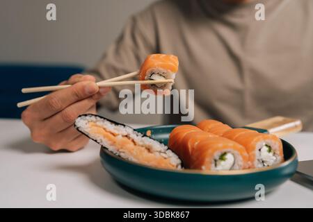 Uomo che usa le bacchette per raccogliere salmone fresco e formaggio spalmabile sushi roll dal piatto di delizioso maki giapponese, gustando un pasto sano e saporito Foto Stock