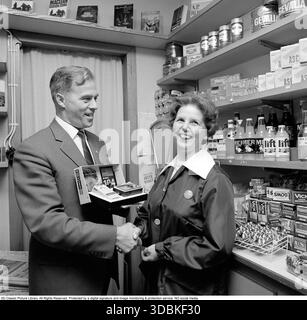 La Kodak Instamatic 104, introdotta nel 1965 e venduta fino alla fine degli anni '1960, era una fotocamera point-and-shoot accessibile e facile da usare che ha contribuito a rendere popolare la fotografia istantanea. Ha utilizzato l'innovativa cartuccia di pellicola 126 di Kodak, che ha consentito un semplice caricamento drop-in, senza la necessità di fastidiose infilature, rendendola ideale per utenti occasionali e famiglie. Con un obiettivo a messa a fuoco fissa, una presa per cubo flash integrato e simboli meteo per le impostazioni di esposizione, la serie Instamatic ha venduto oltre 50 milioni di unità in tutto il mondo, democratizzando la fotografia negli anni sessanta. Nella foto una dipendente donna in un piccolo negozio che riceve un Foto Stock