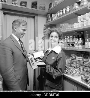 La Kodak Instamatic 104, introdotta nel 1965 e venduta fino alla fine degli anni '1960, era una fotocamera point-and-shoot accessibile e facile da usare che ha contribuito a rendere popolare la fotografia istantanea. Ha utilizzato l'innovativa cartuccia di pellicola 126 di Kodak, che ha consentito un semplice caricamento drop-in, senza la necessità di fastidiose infilature, rendendola ideale per utenti occasionali e famiglie. Con un obiettivo a messa a fuoco fissa, una presa per cubo flash integrato e simboli meteo per le impostazioni di esposizione, la serie Instamatic ha venduto oltre 50 milioni di unità in tutto il mondo, democratizzando la fotografia negli anni sessanta. Nella foto una dipendente donna in un piccolo negozio che riceve un Foto Stock