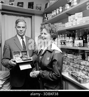 La Kodak Instamatic 104, introdotta nel 1965 e venduta fino alla fine degli anni '1960, era una fotocamera point-and-shoot accessibile e facile da usare che ha contribuito a rendere popolare la fotografia istantanea. Ha utilizzato l'innovativa cartuccia di pellicola 126 di Kodak, che ha consentito un semplice caricamento drop-in, senza la necessità di fastidiose infilature, rendendola ideale per utenti occasionali e famiglie. Con un obiettivo a messa a fuoco fissa, una presa per cubo flash integrato e simboli meteo per le impostazioni di esposizione, la serie Instamatic ha venduto oltre 50 milioni di unità in tutto il mondo, democratizzando la fotografia negli anni sessanta. Nella foto una dipendente donna in un piccolo negozio che riceve un Foto Stock