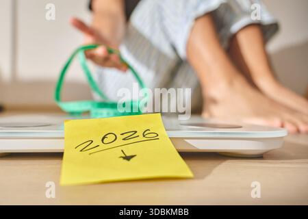 Persona in pigiama seduta su una bilancia con un metro a nastro e una nota di risoluzione 2026 Foto Stock