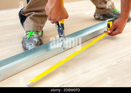 L'operaio edile sta tagliando un profilo metallico con cesoie per aviazione e misura con metro a nastro, durante lavori di ristrutturazione e miglioramento della casa Foto Stock