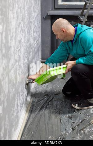 L'immagine mostra un uomo calvo che applica intonaco decorativo a una parete in una stanza. Si sta inginocchiando sul pavimento, che è ricoperto da un telo di plastica per pr Foto Stock