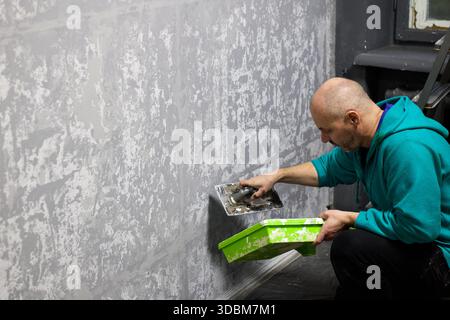 Questa immagine mostra un artigiano che applica intonaco decorativo a una parete utilizzando una cazzuola. Il lavoratore, vestito con una felpa casual con cappuccio, si concentra sul compito di h Foto Stock