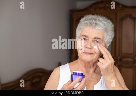 donna anziana di 70 anni che applica crema per il viso. cura di sé, routine di bellezza, anti-invecchiamento e cura della pelle per donne mature Foto Stock