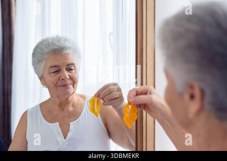 Donna anziana che applica la maschera labiale e controlla il riflesso nello specchio a casa, routine di bellezza, cura della pelle, cura di sé, anti invecchiamento e donna matura sicura di sé Foto Stock