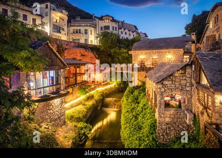 Affascinanti ristoranti illuminati e case in pietra lungo un piccolo fiume nella città vecchia di Mostar al crepuscolo, Mostar, Herzegovina Neretva Canton, Bosnia ed Erzegovina Foto Stock