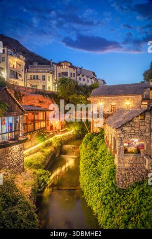 Affascinanti ristoranti illuminati e case in pietra lungo un piccolo fiume nella città vecchia di Mostar al crepuscolo, Mostar, Herzegovina Neretva Canton, Bosnia ed Erzegovina Foto Stock