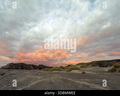 Vista di una spiaggia di sabbia ferma che incontra aspre scogliere sotto un cielo spettacolare che si accendono con sfumature di tramonto sulla spiaggia di Wharariki, Pūponga, regione di Tasman, nuova Zelanda Foto Stock