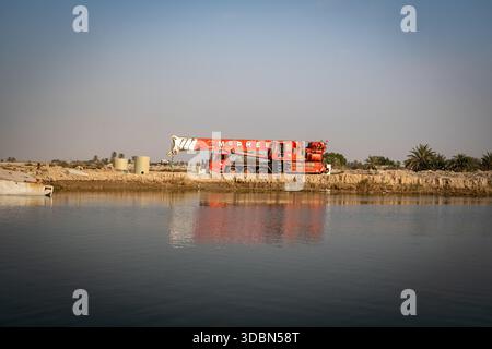 Grande gru mobile rossa parcheggiata sulla Earthen River Bank vicino a detriti sommersi e palme, che rappresentano lo sviluppo industriale. Foto Stock