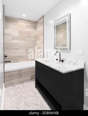 Un bagno moderno presenta una vasca circondata da piastrelle di legno marrone e un ripiano in marmo con un rubinetto nero su un mobile nero con piastrelle a chevron Foto Stock