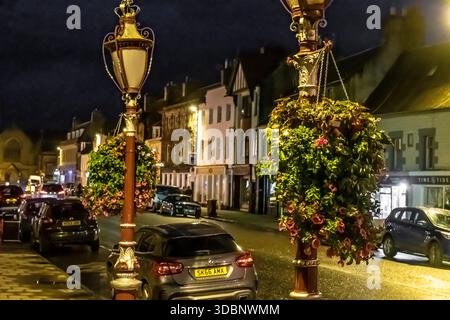 Notte su Peebles High Street con lampade ornate, cesti di fiori appesi e auto parcheggiate lungo edifici storici. Foto Stock