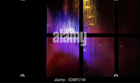 Una finestra scura con gocce di pioggia di notte. Luci natalizie gialle e blu sfocate attraverso il vetro bagnato ricoperto di gocce di condensa. Foto Stock
