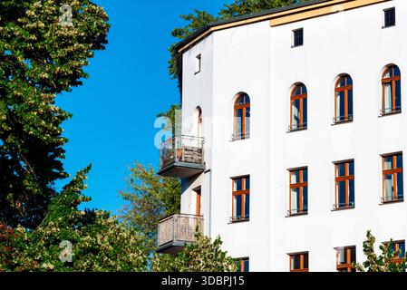 Facciata bianca rinnovata di appartamenti con finestre ad arco in legno e balconi circondati da lussureggianti alberi verdi a Moritzplatz, Berlino. Città soleggiata Foto Stock