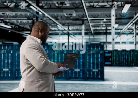 Amministratore che cammina nel data center eseguendo linee di codice sul notebook, risolvendo i problemi dei montaggi su rack. Esperto DI IT afroamericano che utilizza script di programmazione su un notebook in sala server Foto Stock