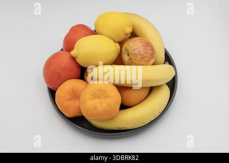 Frutta fresca assortita in una ciotola nera con banane, limoni, arance, mele e cachi isolati su sfondo bianco Foto Stock