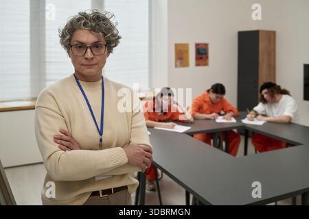 Donna caucasica di mezza età con capelli corti ricci in piedi con le braccia incrociate in primo piano, tre giovani donne adulte in uniforme arancione sedute a tavoli a studiare in aula Foto Stock