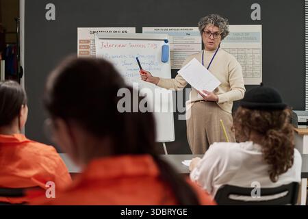 Donna caucasica di mezza età che insegna un gruppo di giovani donne adulte in uniforme arancione, scrive su lavagna con consigli per colloqui di lavoro, gli studenti ascoltano attentamente in aula Foto Stock