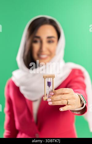 "Una donna con un velo bianco e un blazer rosa regge una piccola clessidra di legno con sabbia viola su un solido sfondo verde. Foto Stock
